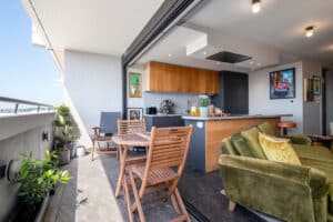 Modern apartment with open kitchen and living area leading to a balcony. Wooden dining set, green sofa, and plants decorate the space.