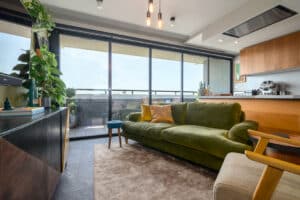 Modern living room with large windows, green velvet sofa, wooden furniture, and plants. Kitchen area visible in the background. Bright and stylish interior.