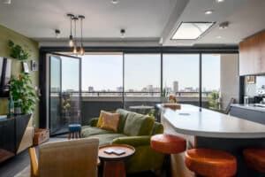 Modern living room with large windows opening to a city view, featuring a green sofa, wooden accents, and a kitchen area with a white countertop.