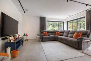 Modern living room with a large gray sectional sofa, orange pillows, a flat-screen TV on a blue console, and large windows overlooking greenery.