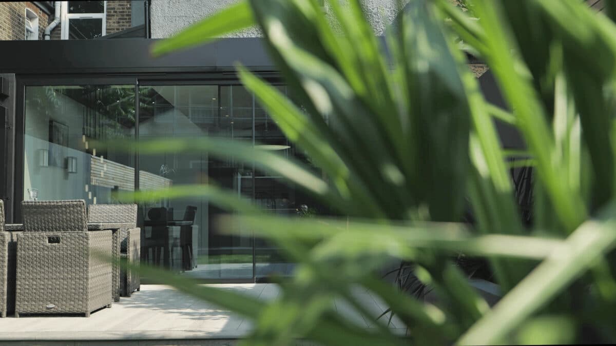 A modern outdoor patio featuring wicker furniture is partly hidden by lush green leaves, seamlessly connected to the interior space through elegant glass sliding doors.