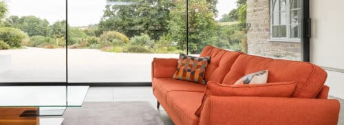 A modern living room with a large window showing a garden view. It features an orange sofa with patterned cushions, a glass coffee table, and a stone wall.