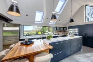 Modern kitchen featuring aluminium casement windows that flood the space with natural light, dark cabinets, a white island, and a wooden dining table with beige chairs. Skylights enhance the open feel while large windows offer serene views of the surrounding greenery.