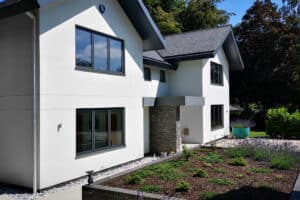 Modern white two-story house with large aluminium casement windows, a gray roof, and stone accents. It features a landscaped front yard with a small garden and greenery.