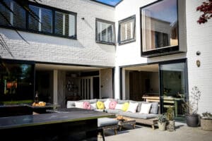 Modern patio with white brick walls, outdoor seating adorned with colorful cushions, and sleek aluminium casement windows beside sliding glass doors leading to an open-plan interior.