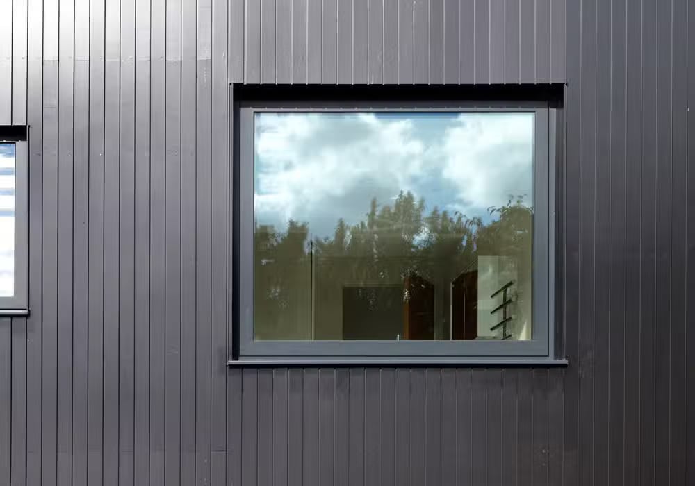 An aluminium casement window on a dark gray paneled wall reflects a cloudy sky and trees.