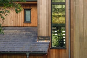 Exterior of a building with a slate roof, wooden siding, and two aluminium casement windows, one narrow and one large, reflecting greenery.