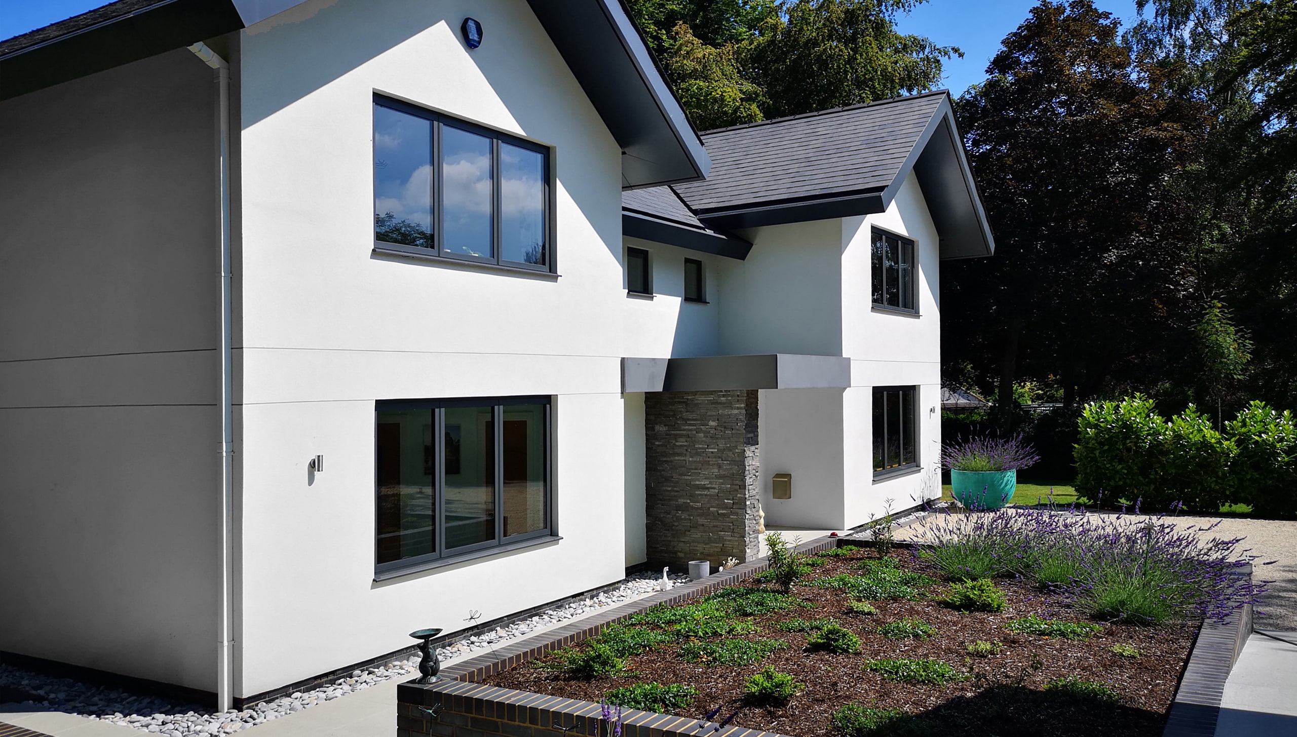 A modern two-story house with white walls, dark roof, and stone accent features sleek aluminium casement windows. It is surrounded by a garden with greenery and purple flowers. The sky is blue with scattered clouds.