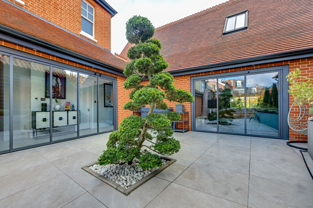 Courtyard with a sculpted evergreen tree on a pebble base, surrounded by brick walls and large slide and turn glass doors on a tiled floor.