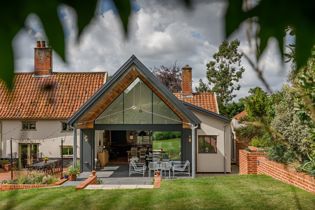 A modern house with a glass-fronted extension and gable window features an outdoor dining area and a red tiled roof, all set in a garden adorned with trees and encircled by a brick wall.