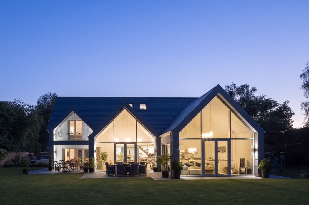 Modern house with large glass windows, including a striking gable window, glowing brightly in the evening, set against a clear blue sky.