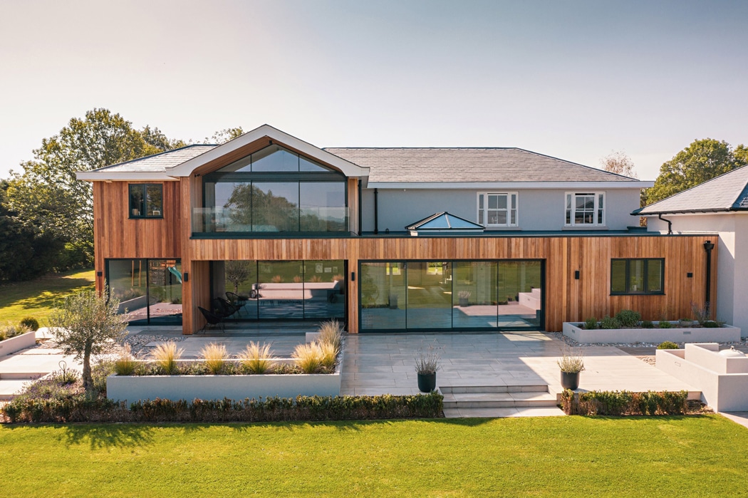 Modern two-story house with wooden paneling, large glass windows, and a slate roof featuring a charming gable window. The spacious patio is surrounded by a well-manicured lawn and trees, completing this picturesque setting.