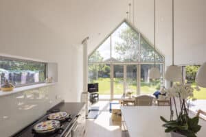 Bright kitchen with vaulted ceiling and a charming gable window, large glass doors, and windows. A dining table, plants, and cooking area with decorative plates on the stove. Garden view visible outside.