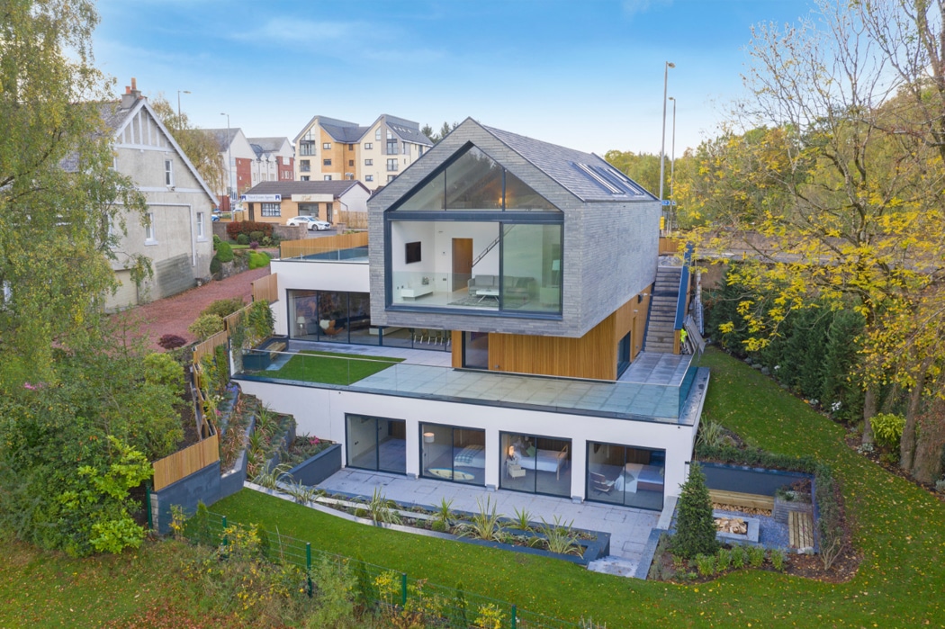 Modern glass-fronted house with three levels, angular roof, and landscaped garden. Large gable windows overlook lush greenery. Adjacent to a residential area with trees and houses in the background.