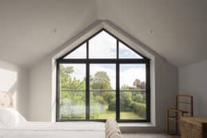 The bedroom features a large gable window overlooking a garden. A bed with a white comforter is accompanied by a wicker basket and wooden shelf, creating a serene and inviting space.