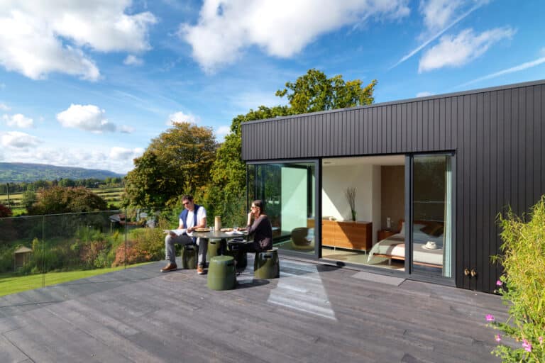 Two people sit at a small table on a modern rooftop patio with glass railing, surrounded by greenery and overlooking a scenic landscape with blue sky and clouds.