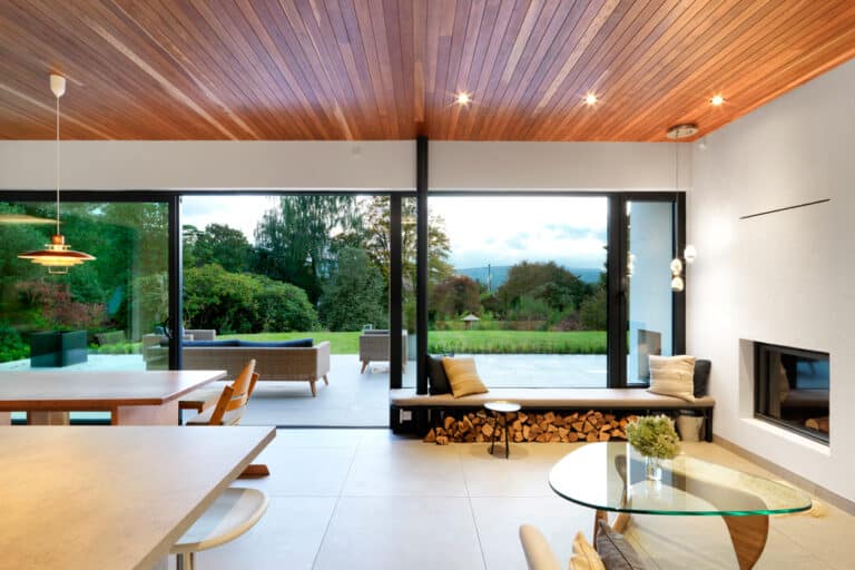 Modern living space with wood ceiling, large windows, and a view of a garden. Features a glass table, a built-in fireplace, and a bench with firewood storage.