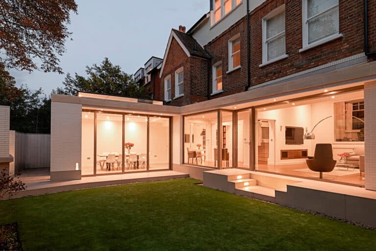 Exterior view of a modern house with large glass doors, illuminated interior, and a manicured lawn in the foreground.