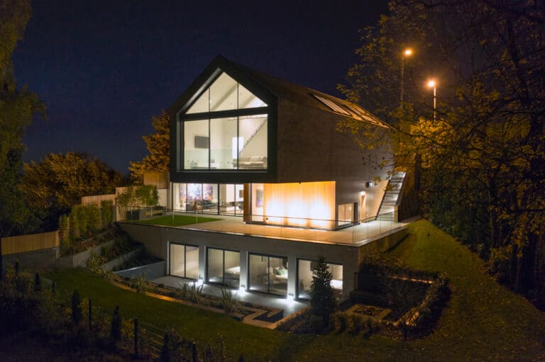 Modern, multi-story house with large glass windows and illuminated interiors, set on a landscaped slope at night.