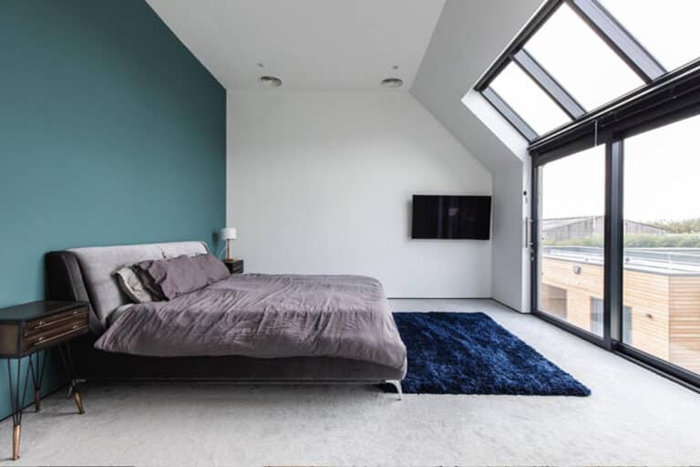 A bedroom with a large bed, blue accent wall, mounted TV, and skylight windows. A navy blue rug is on the light-colored floor.