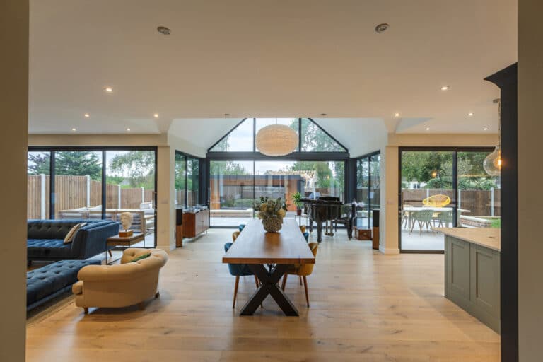 Spacious open-plan living room with a wooden dining table, blue sofas, a grand piano, and large glass doors leading to an outdoor area.