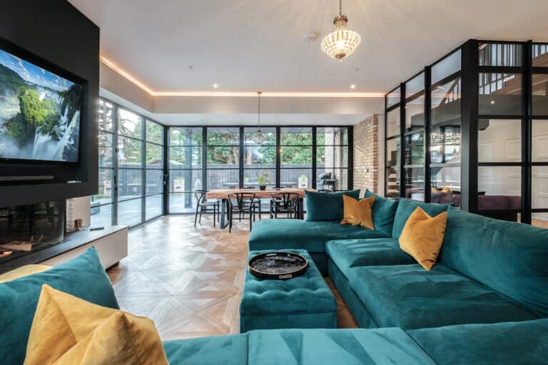 Spacious modern living room with a teal sectional sofa, yellow pillows, large windows, a TV, and a chandelier. Glass partition separates the dining area with a long wooden table.