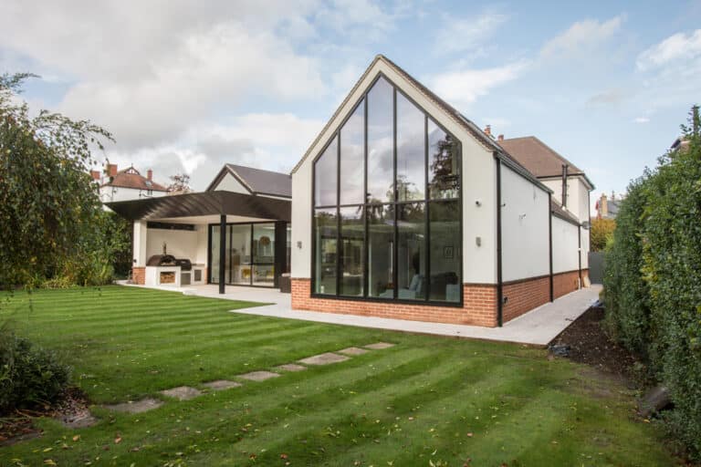 Modern white house with large glass windows, peaked roof, and brick accents. The house is surrounded by a neatly manicured lawn and hedges under a partly cloudy sky.