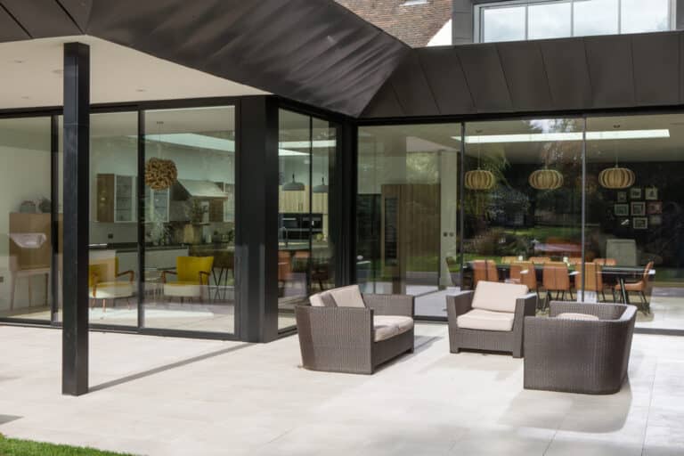 Outdoor patio space with wicker furniture including a sofa and two chairs. Large glass doors open to a modern interior with a kitchen and dining area, featuring pendant lights and yellow accents.