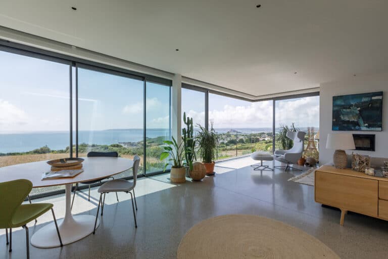 Modern living room with large glass windows offering an ocean view. It features a dining table, chairs, plants, and contemporary decor.