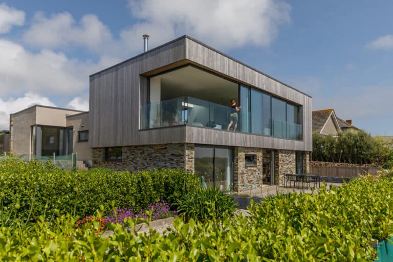 Modern two-story house with large glass balcony, surrounded by greenery. A person stands on the balcony looking outward. The exterior features wood and stone accents.