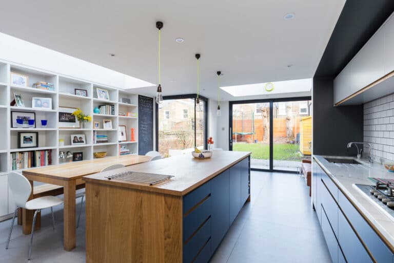 Modern kitchen with a wooden island, blue cabinets, dining area, bookshelves on the left, and large windows opening to a garden. Three pendant lights hang above the island.