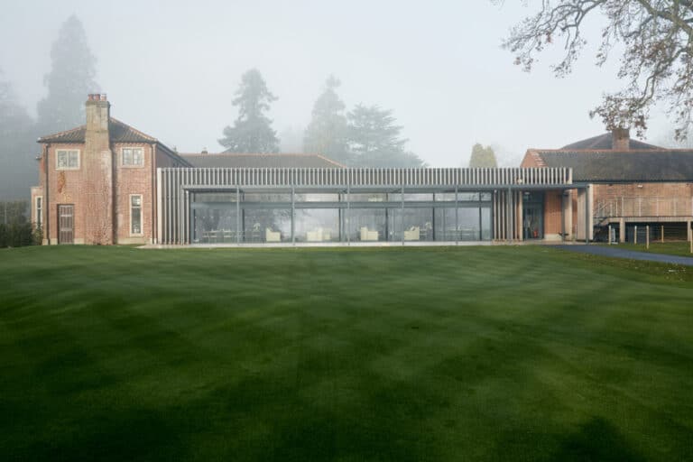 A modern glass extension on a traditional brick building, surrounded by a lush green lawn and trees in the background on a misty day.