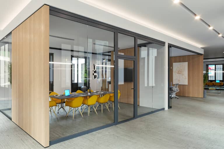 Modern office space with glass-walled meeting room. Inside, a long table with yellow chairs, a mounted screen, and a laptop. Open area outside with seating and ceiling lights.