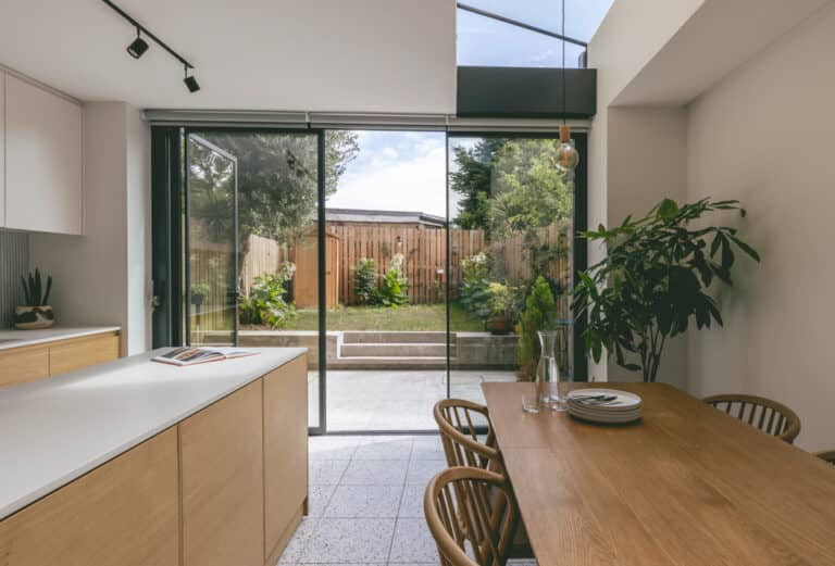 Modern kitchen and dining area with wooden furniture, large glass doors opening to a fenced garden, and potted plants.