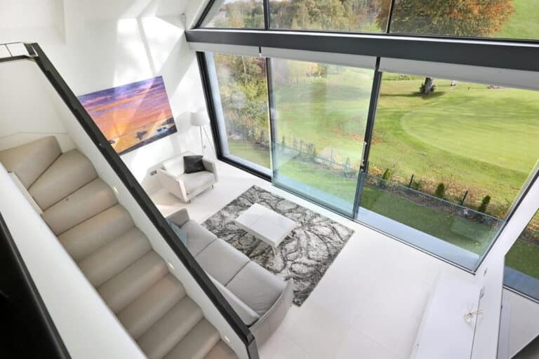 Modern living room with large windows overlooking a golf course. Features a staircase, beige sofa, abstract wall art, and a gray rug on white tile floor.