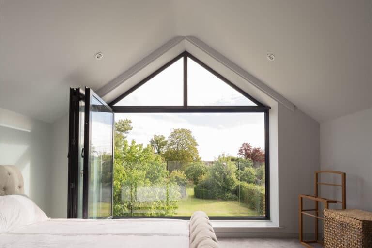 A bedroom with a large triangular window overlooks a lush green garden. The window is partially open, and there is a wicker basket and chair in the corner.