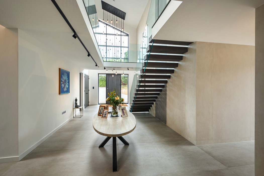 Modern interior with a circular table displaying flowers and frames. A staircase with glass railings leads to an upper floor, where a gable window adds charm and light streams through, illuminating the space beautifully.