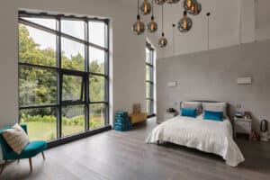 Modern bedroom with a gable window, large double bed, teal accents, and hanging pendant lights. A chair and stacked boxes sit in the corner, overlooking a lush outdoor view.