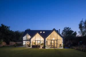 The modern house features expansive glass and a striking gable window, illuminated from within against a dark blue evening sky, nestled among trees and a lush grassy lawn.