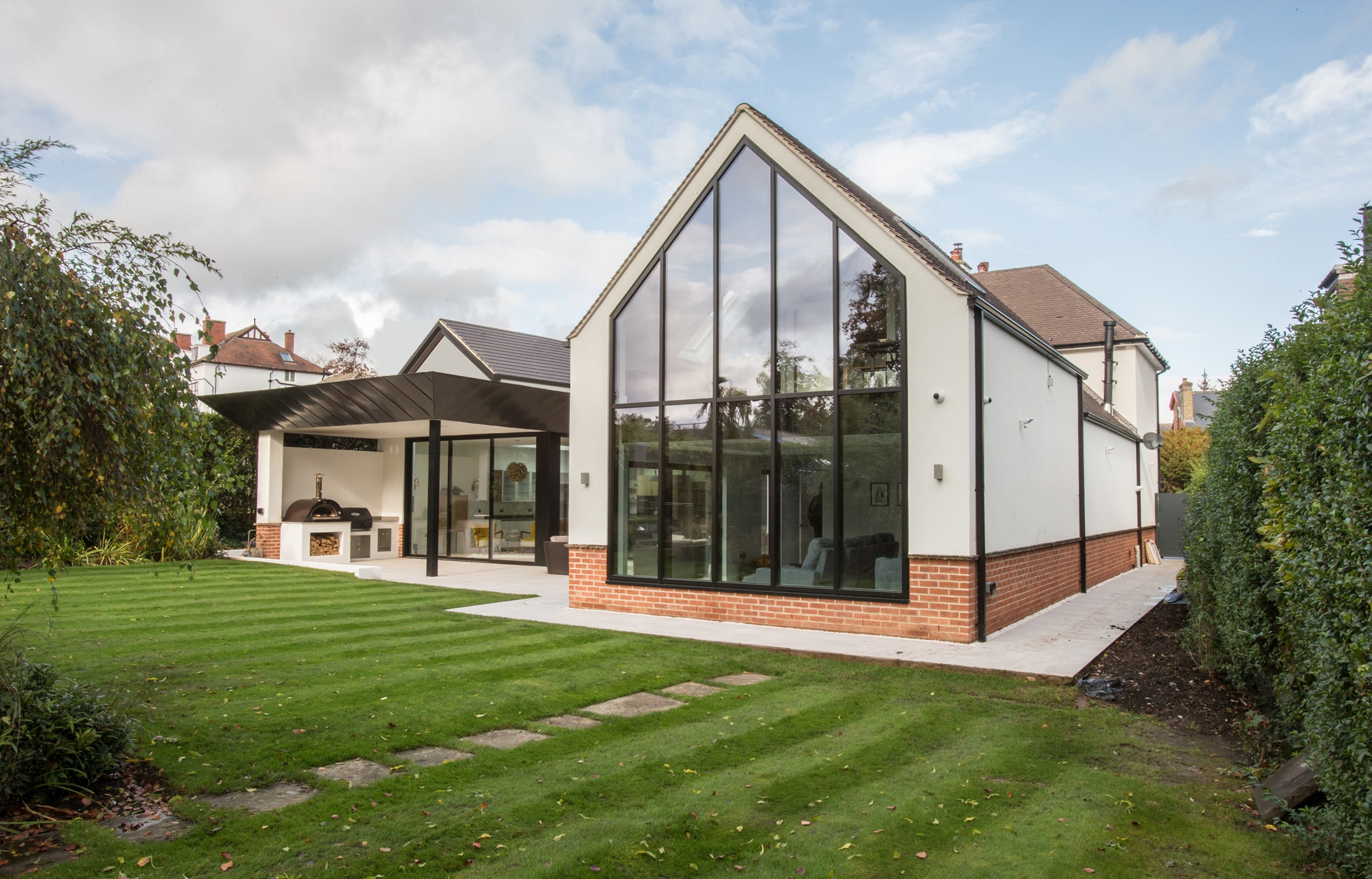 Modern house with expansive glass and gable windows, complemented by a sloped roof. It sits adjacent to a well-manicured lawn bordered by a hedge. Enjoy the patio area with stylish outdoor furniture, surrounded by elegant green trees.