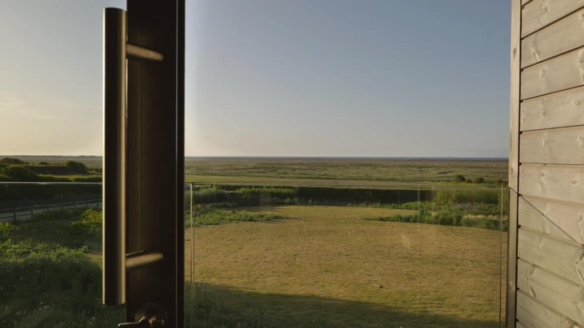 A frameless glass balustrade surrounds the open window, offering an unobstructed view of the grassy landscape and distant ocean, all bathed in the warm hues of a clear sunset sky.