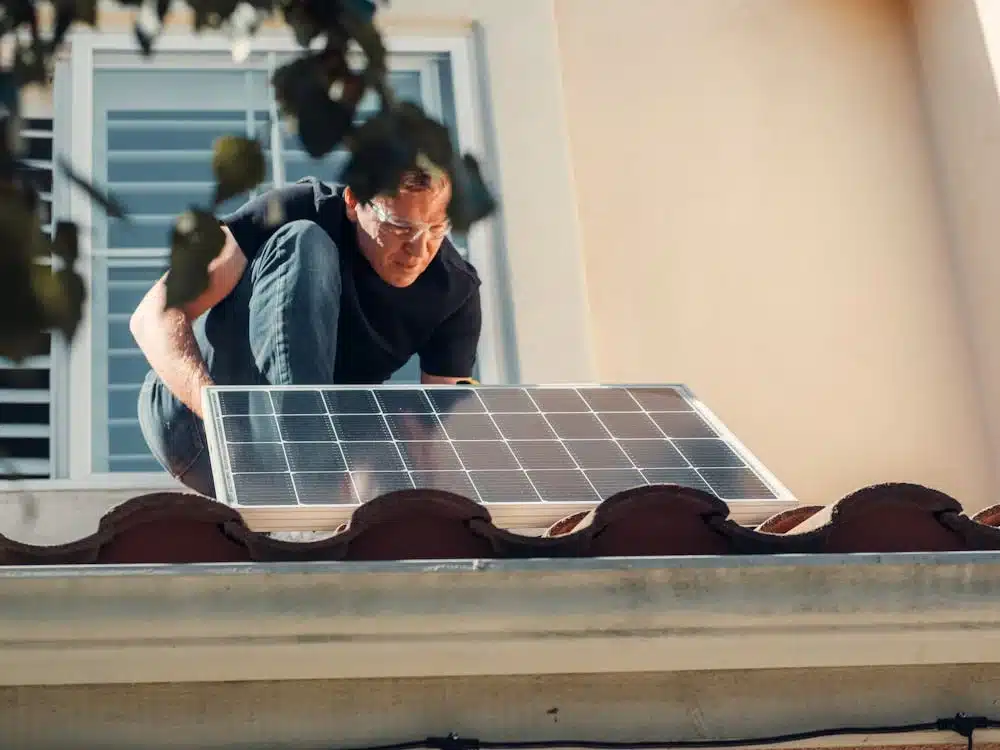 Credit: Kindel Media A person installs a solar panel on a rooftop, wearing safety goggles and crouching near a window.