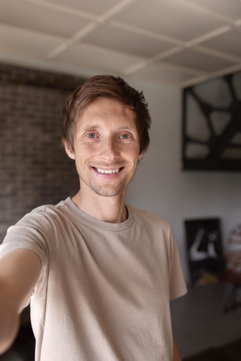 Person wearing a beige t-shirt smiles while taking a selfie indoors.