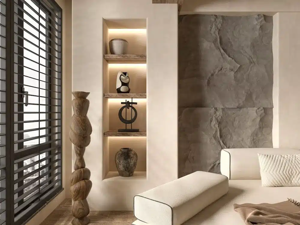 Credit: Wilcle Nunes A modern living room with natural tones features recessed shelves displaying vases and sculptures, a textured wall panel, a large wooden sculpture, and a window with horizontal blinds.