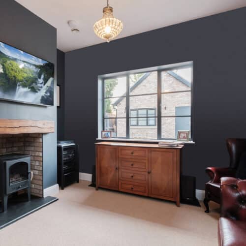 A cozy living room with a dark wall, fireplace, wooden cabinet, armchair, and large window. A landscape photo is mounted above the fireplace.