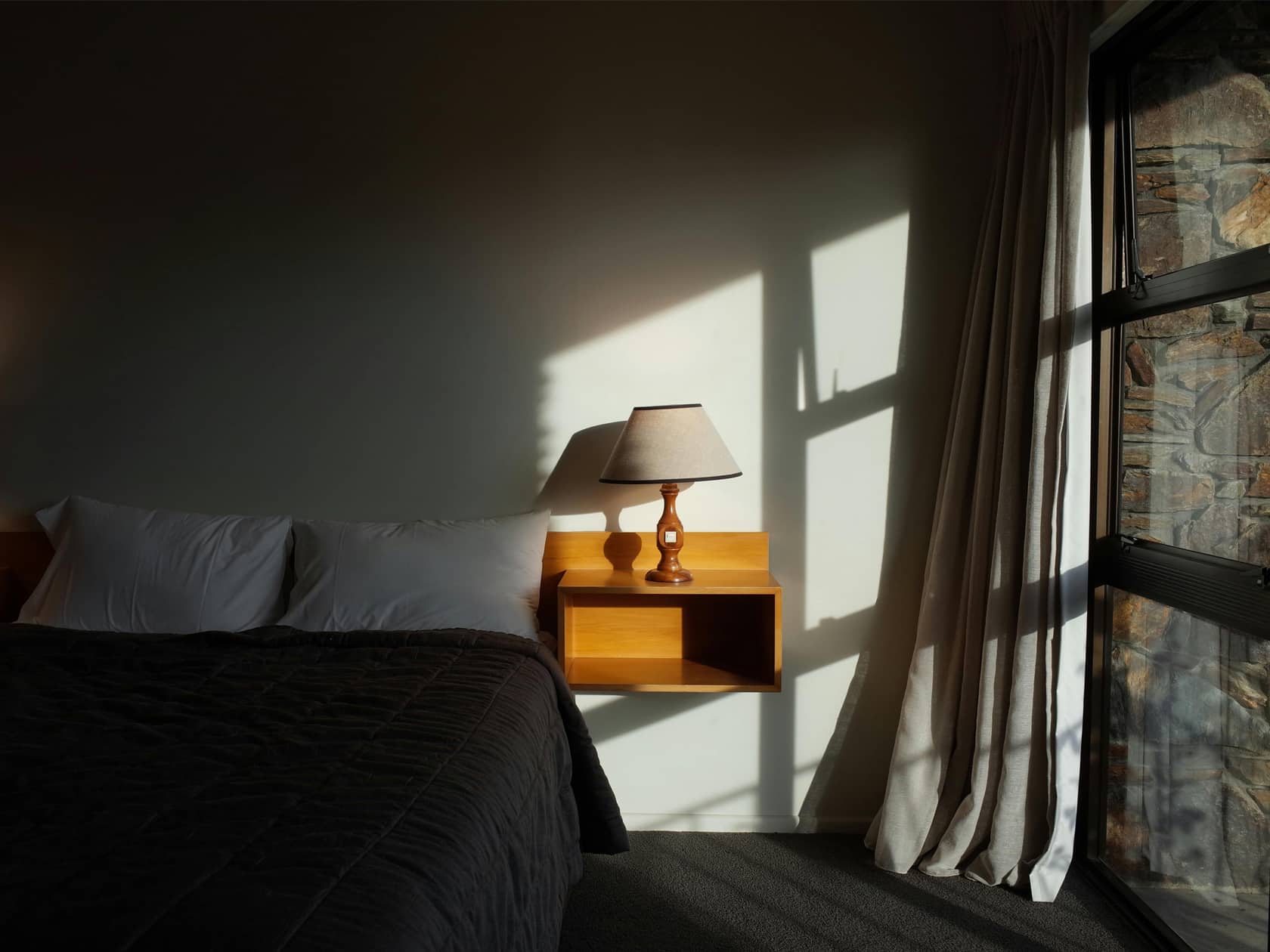 Image Credit: Mingche Lee / A modern bedroom with a black bedspread, a wooden floating shelf and a lamp, accentuated by natural shadows and soft natural light from a large window.