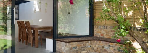 Modern home extension with large black-framed sliding glass doors opening to a patio with a built-in brick bench, surrounded by greenery and red flowering plants