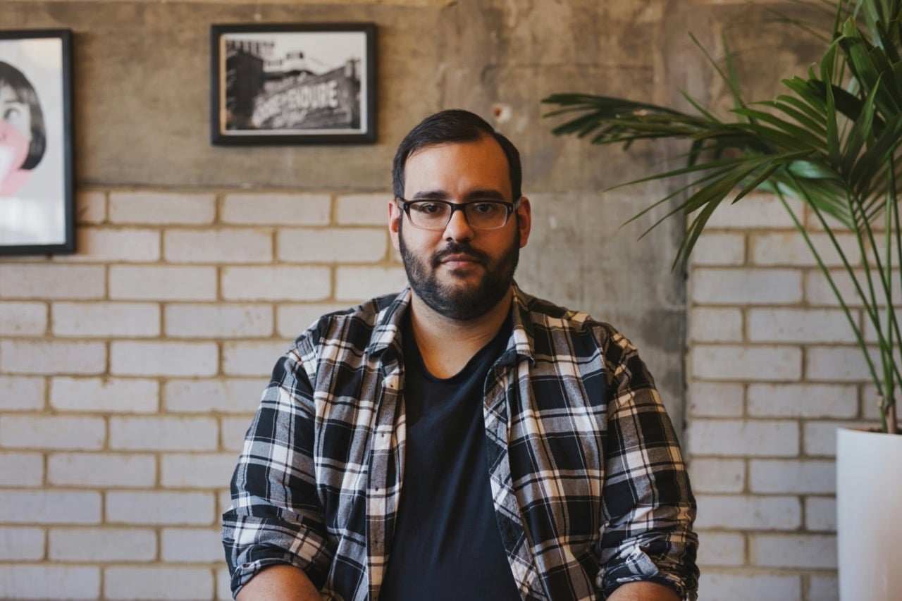 A man with glasses and a beard sits in front of a brick and concrete wall, wearing a black and white plaid shirt. There are two framed pictures and a plant in the background.