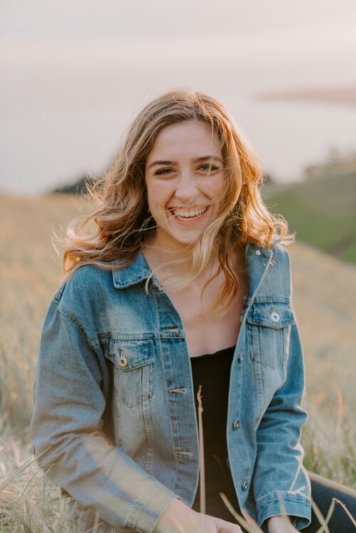 A person with long hair smiles in a grassy field, wearing a denim jacket and black top. The background shows blurred hills and a body of water under a clear sky.