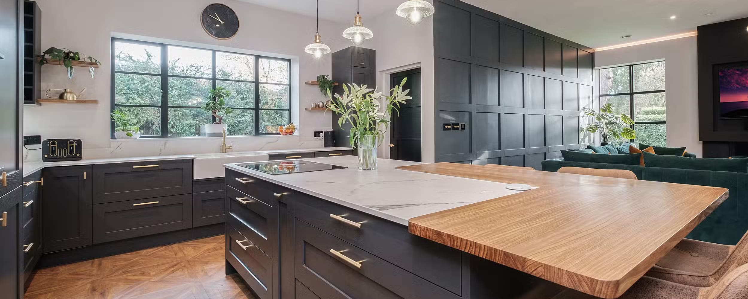 Modern kitchen with black cabinets, marble-topped island with wood extension, pendant lights, and large aluminium windows. A green sofa and plants add a touch of nature to the open-plan space.